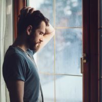 sad man touching his head while standing near window