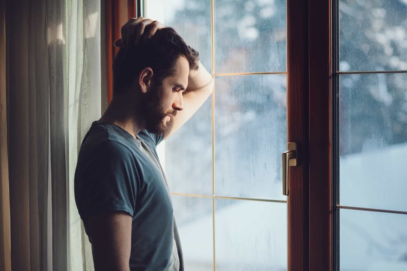 sad man touching his head while standing near window