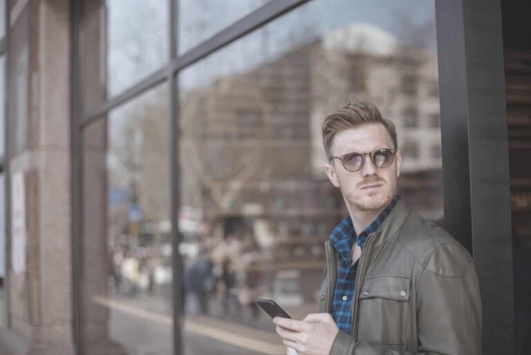 pensive young man looking away and leaning on wall holding a mobile phone