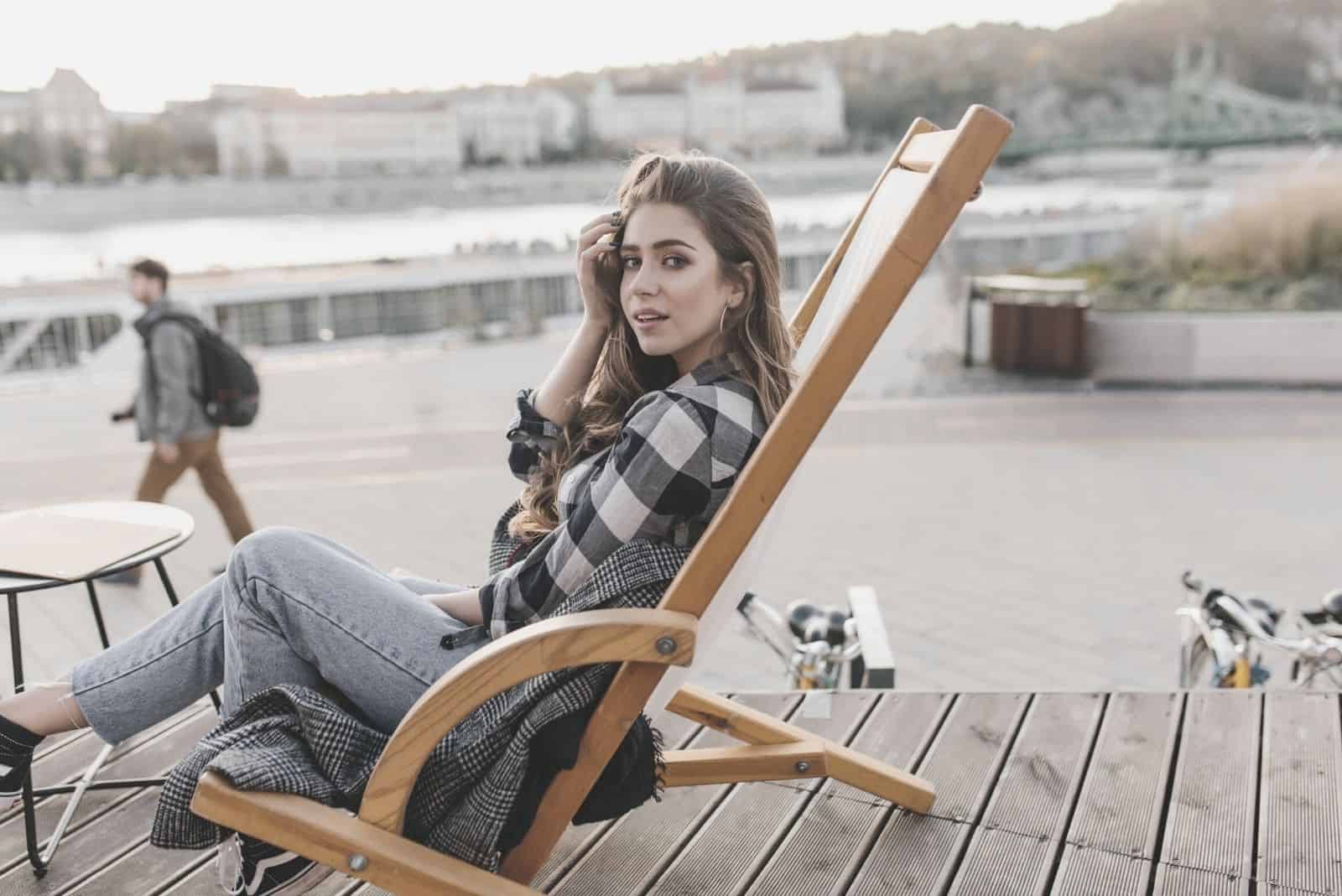 attractive woman wears checkered top sitting on the chair by the wooden platform near the body of water