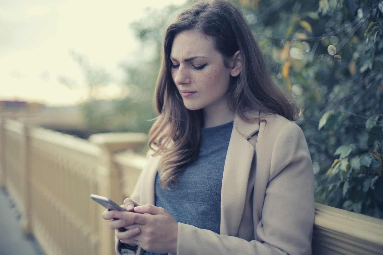mujer pensativa escribiendo un mensaje en su smartphone al aire libre cerca de una valla