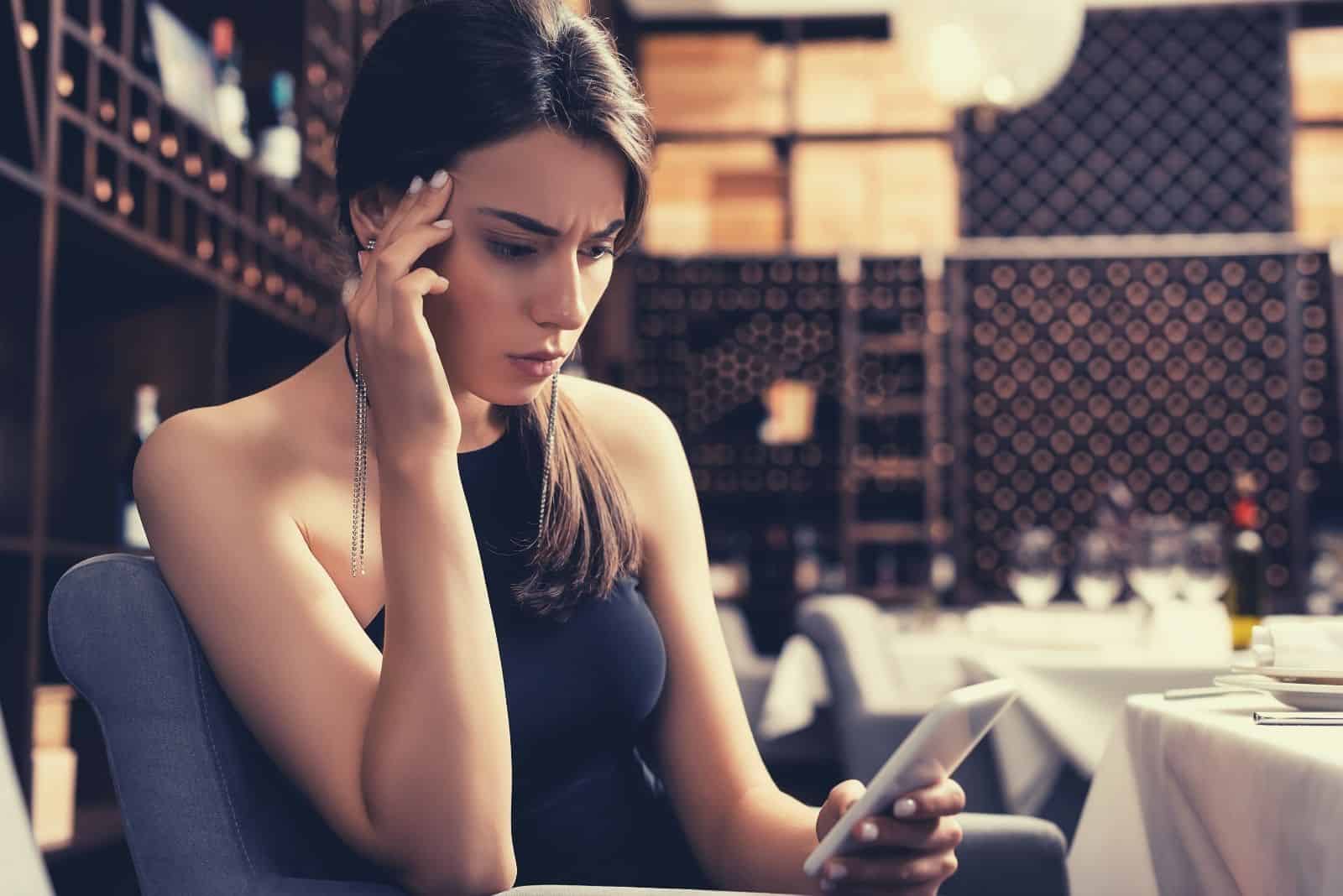 Enfoque selectivo de una mujer pensativa sentada en un restaurante que mira confusa su teléfono móvil.