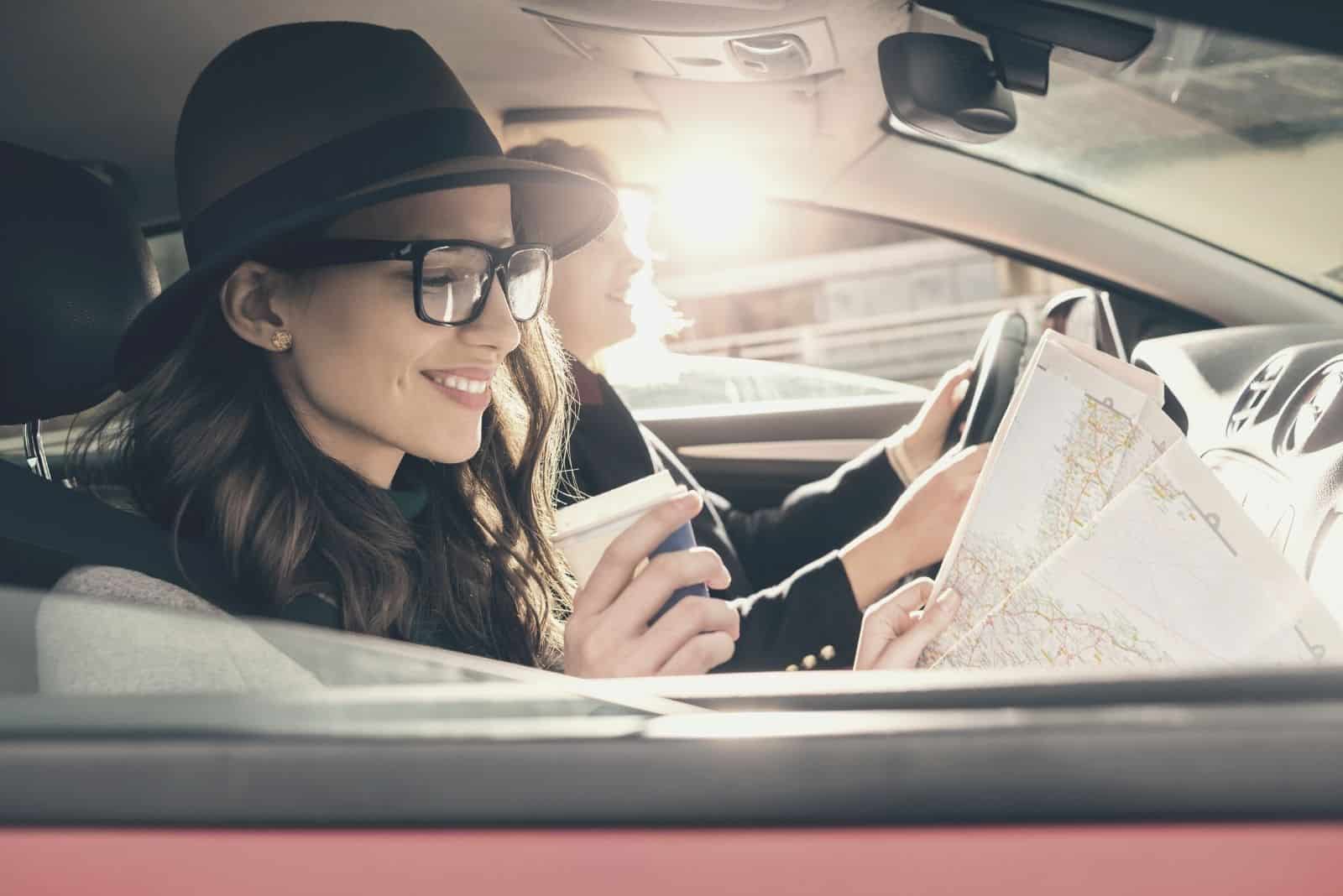 two female friends enjoying their road trip inside the car one looking at the map and a coffee