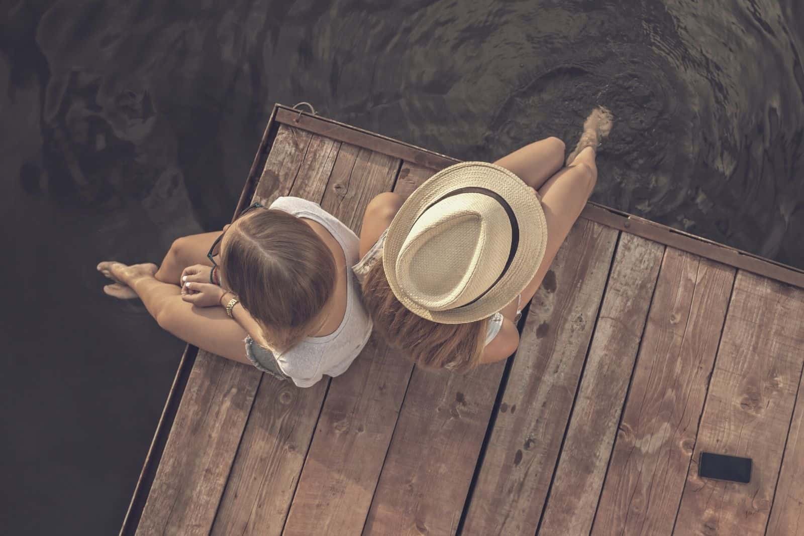 two friends sitting in the wooden platform by the waters in a top angle