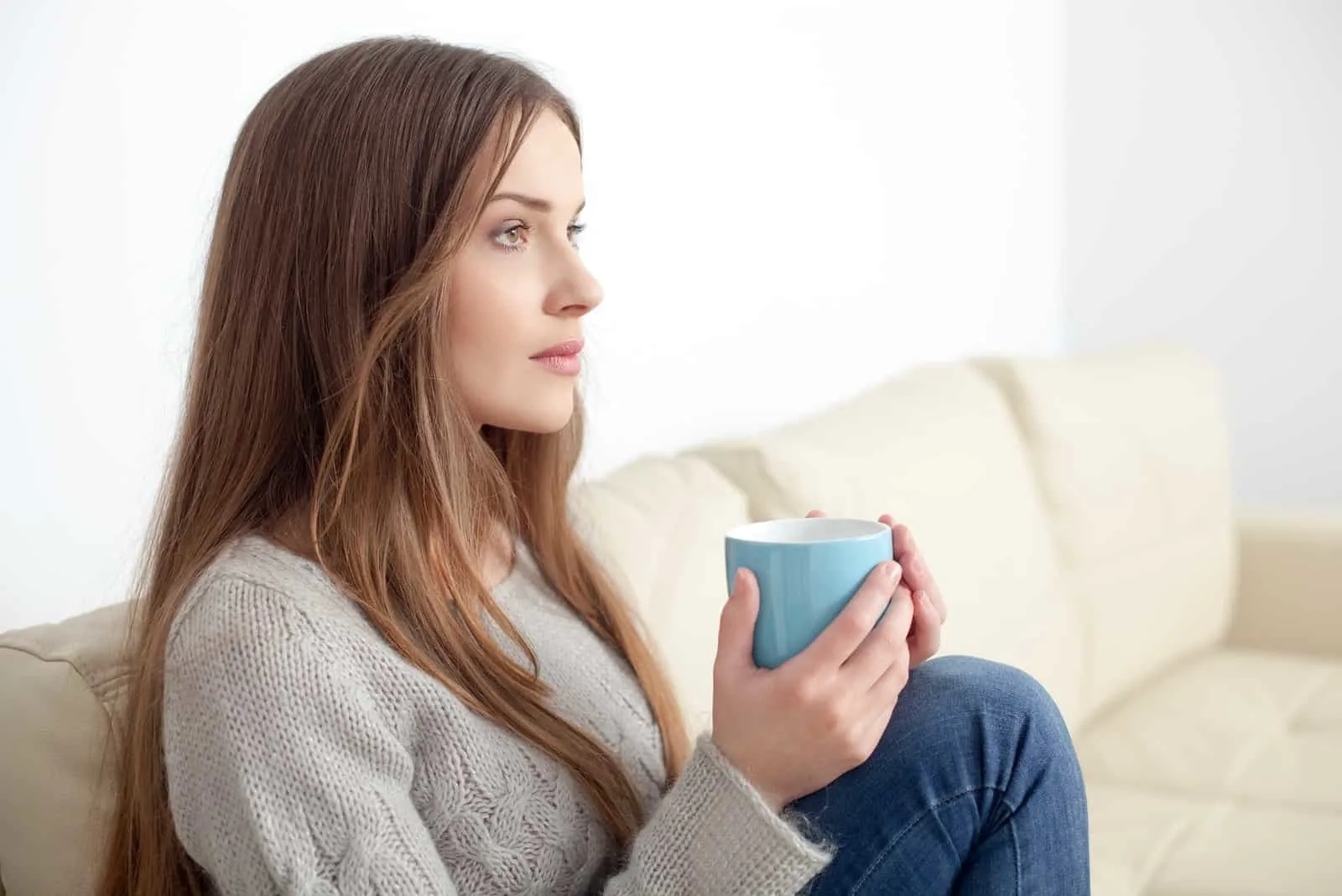 pensive woman holding mug while sitting on sofa