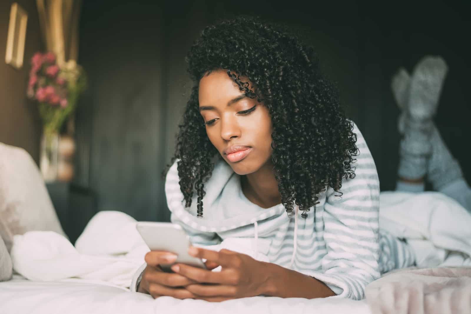 woman with curly hair using smartphone while laying on bed