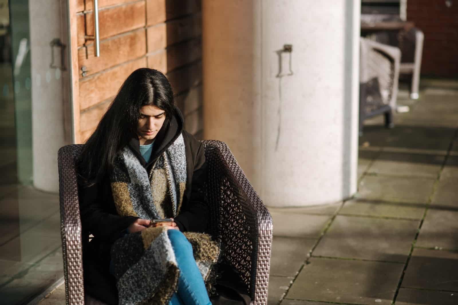 donna con sciarpa che usa lo smartphone mentre è seduta su una sedia