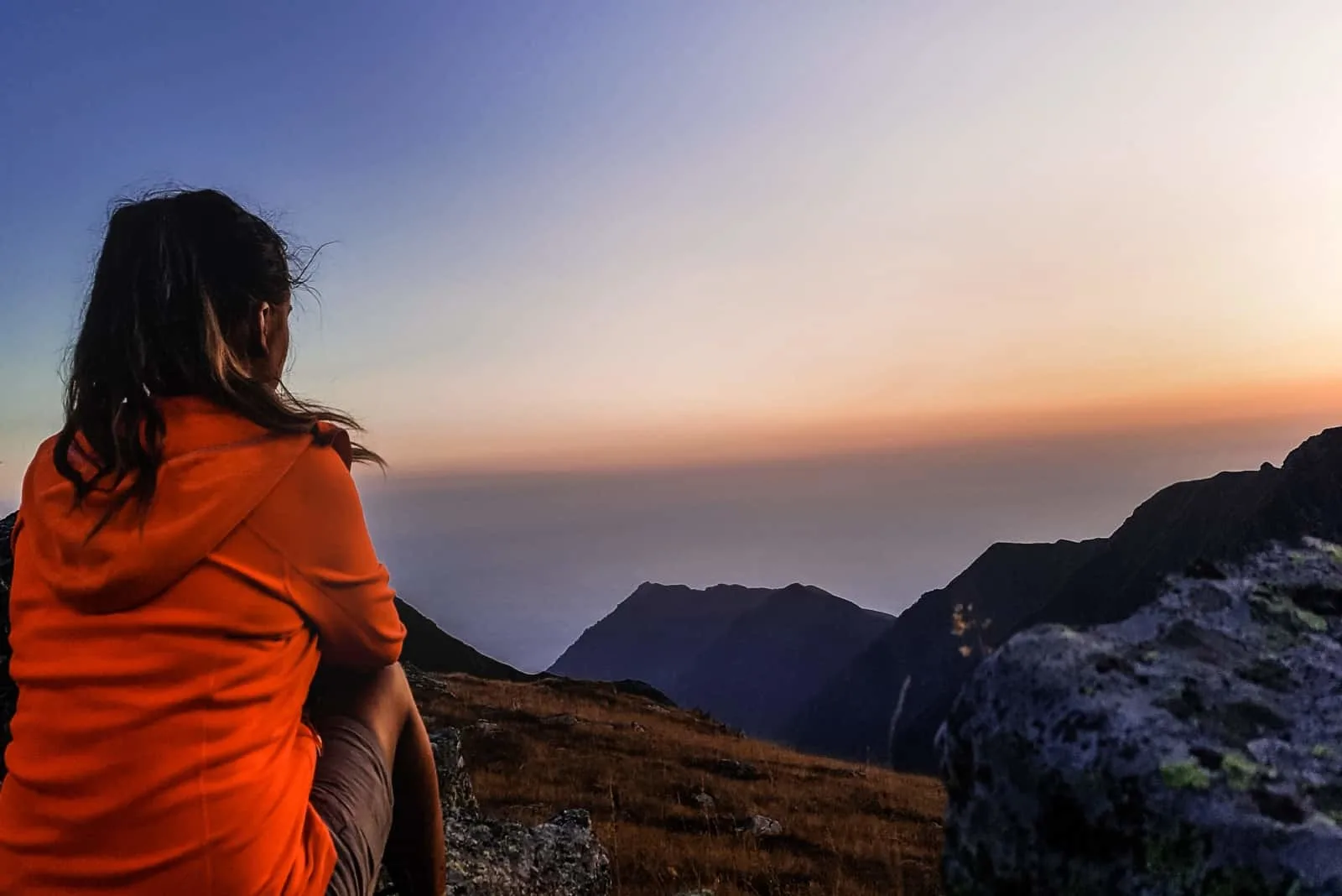 woman in orange jacket sitting on rock looking at mountain