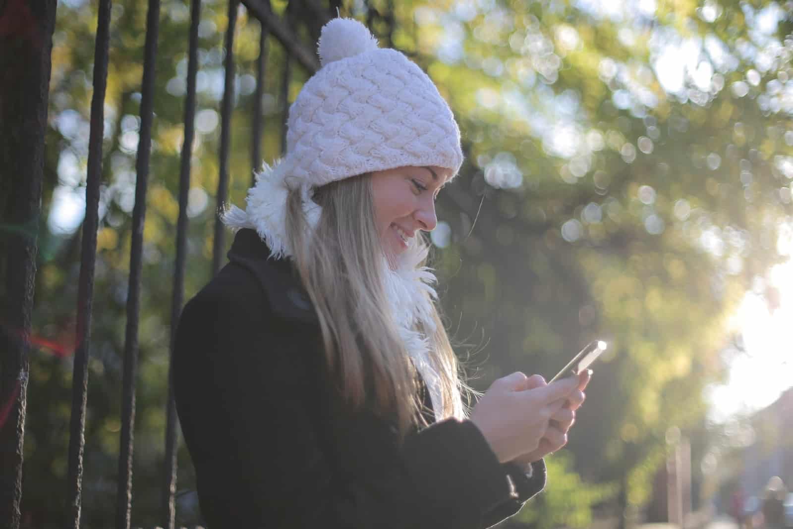 woman with white cap smiling while using smartphone