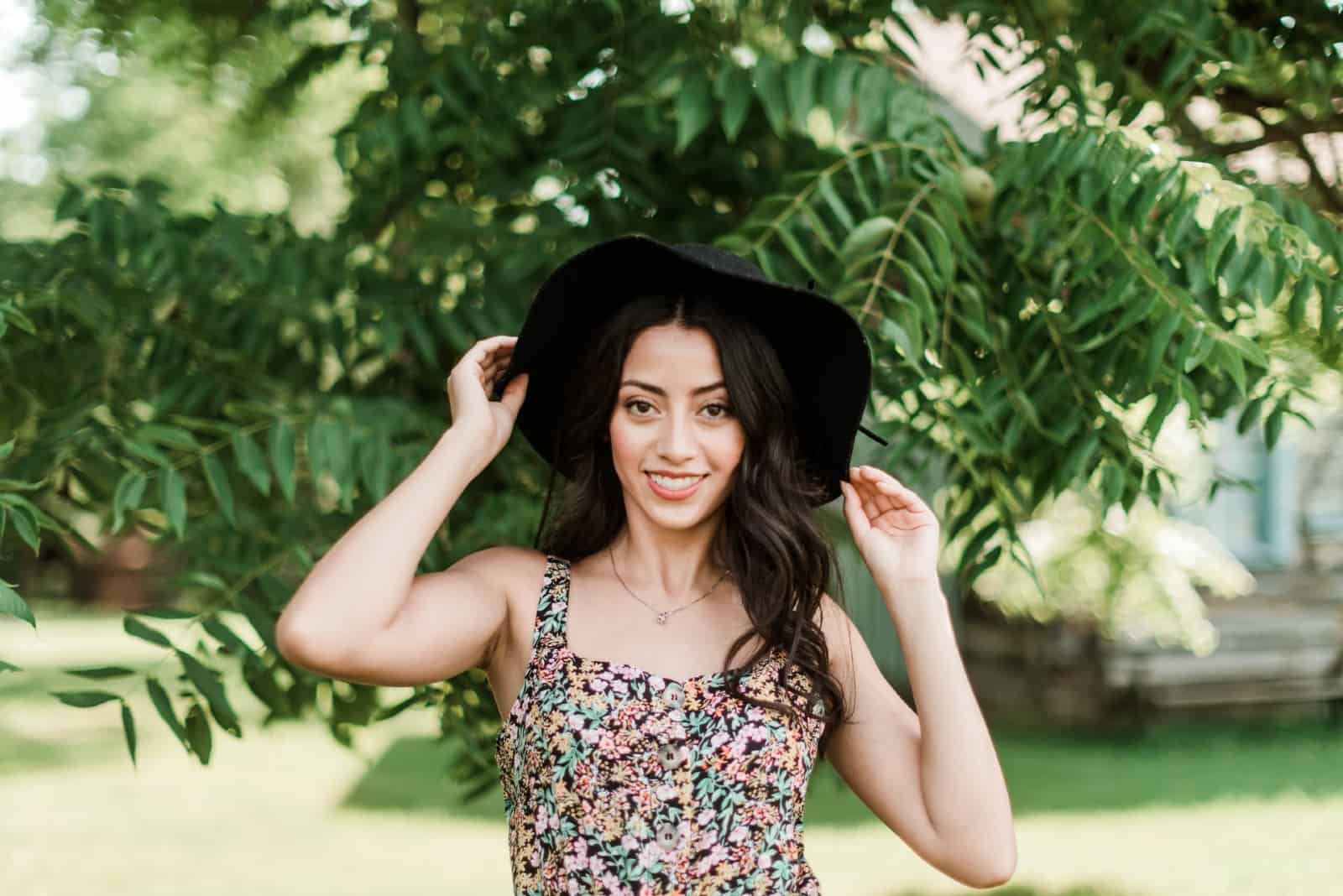 happy woman touching her hat while standing near tree