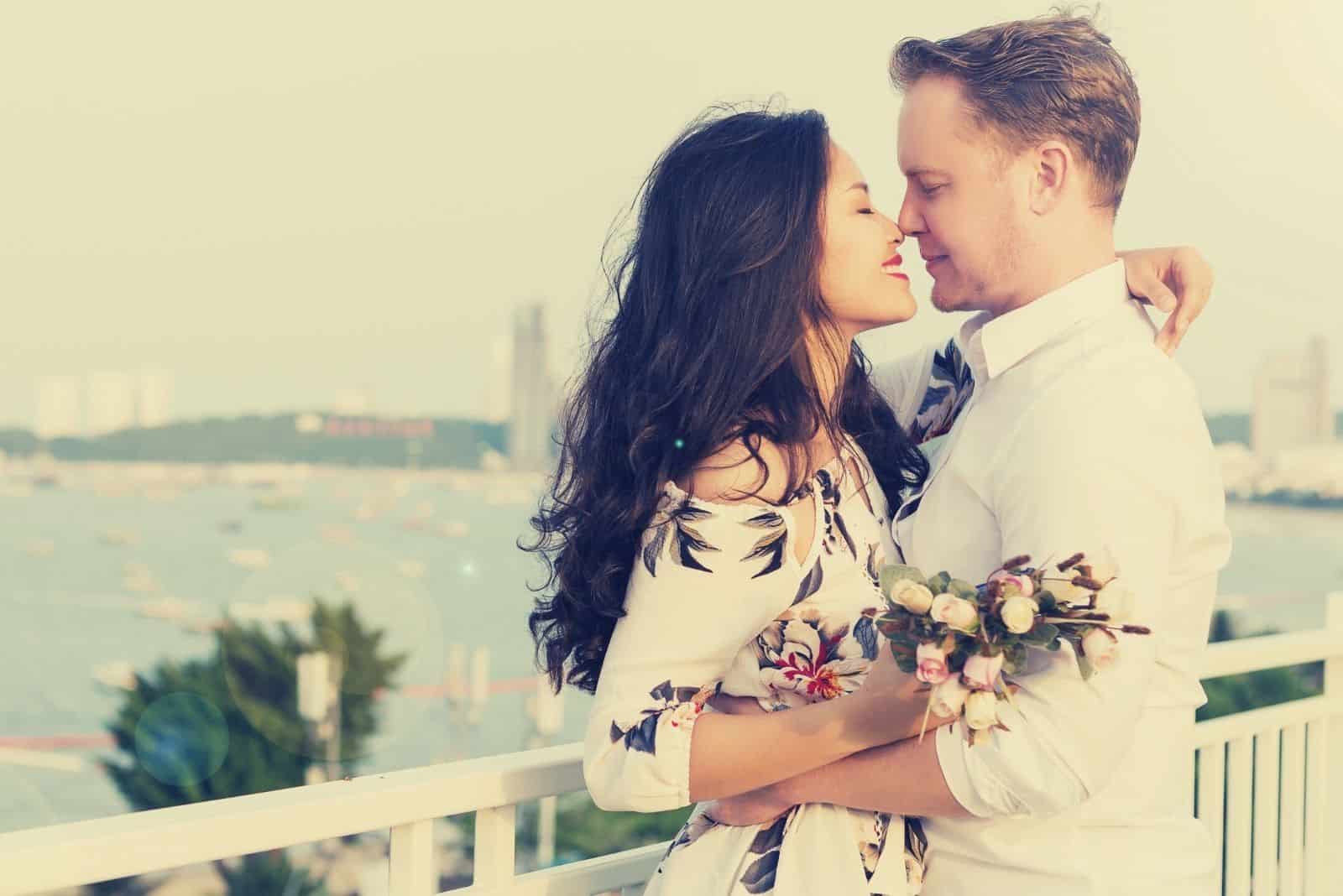 young happy interracial couple hugging and holding a bouquet of flowers near the body of water