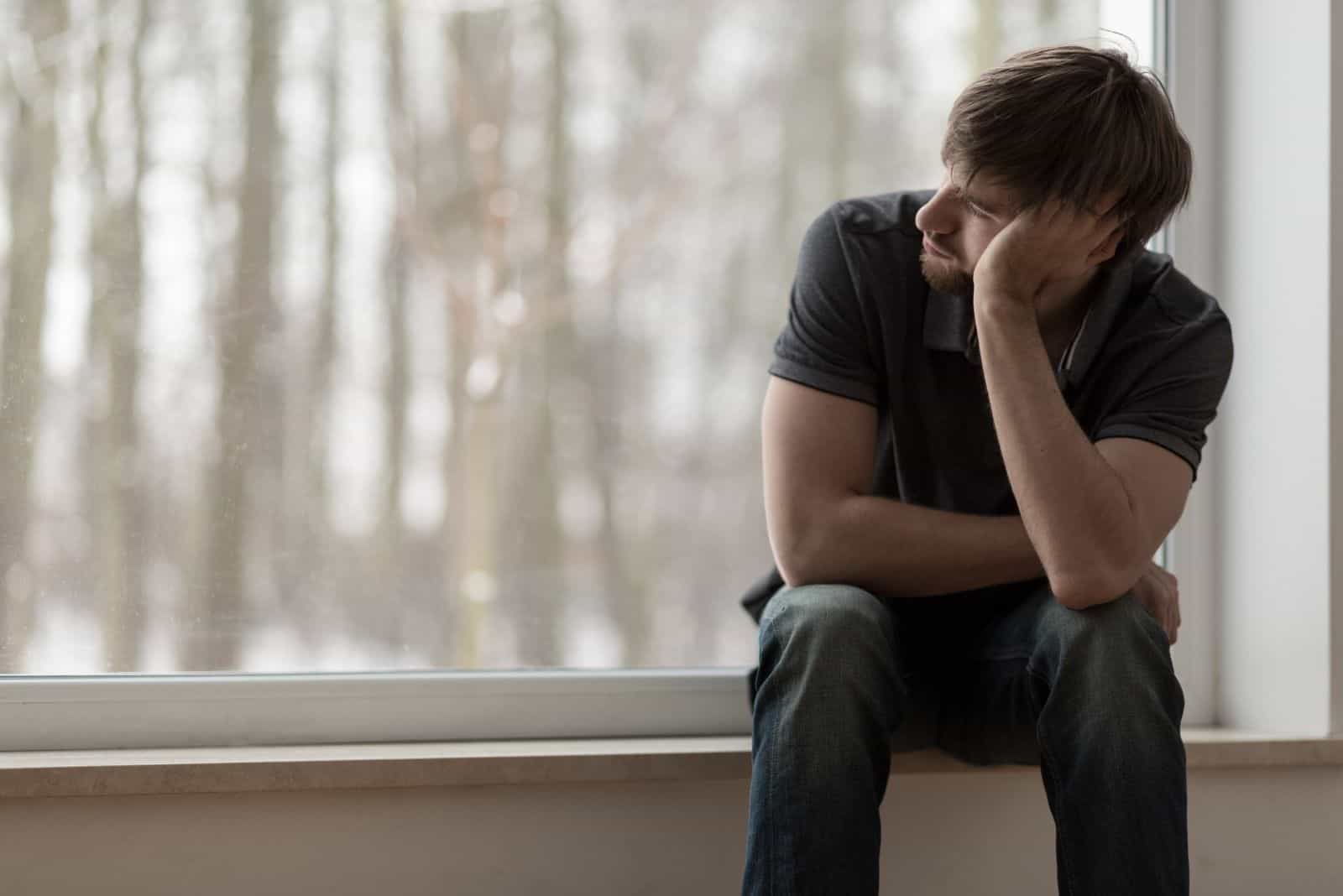 young miserable man sitting by the window sill with trees as the background