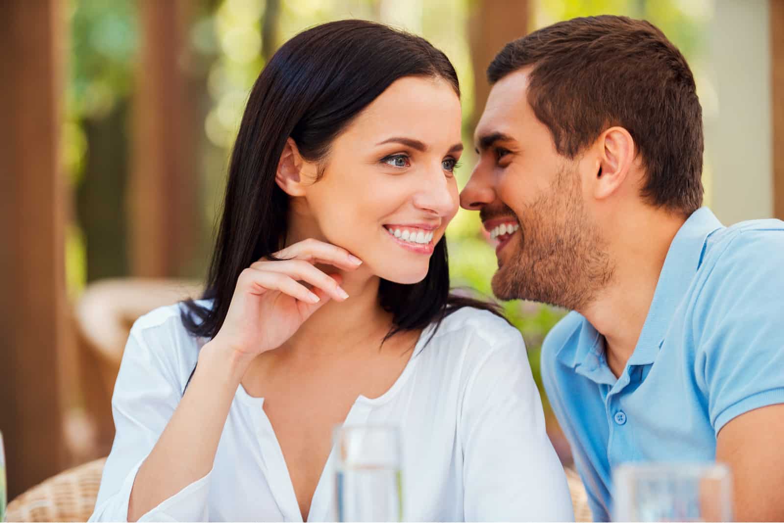 Joven guapo diciéndole algo a su novia y sonriendo mientras están sentados juntos en la mesa al aire libre