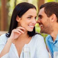 Joven guapo diciéndole algo a su novia y sonriendo mientras están sentados juntos en la mesa al aire libre