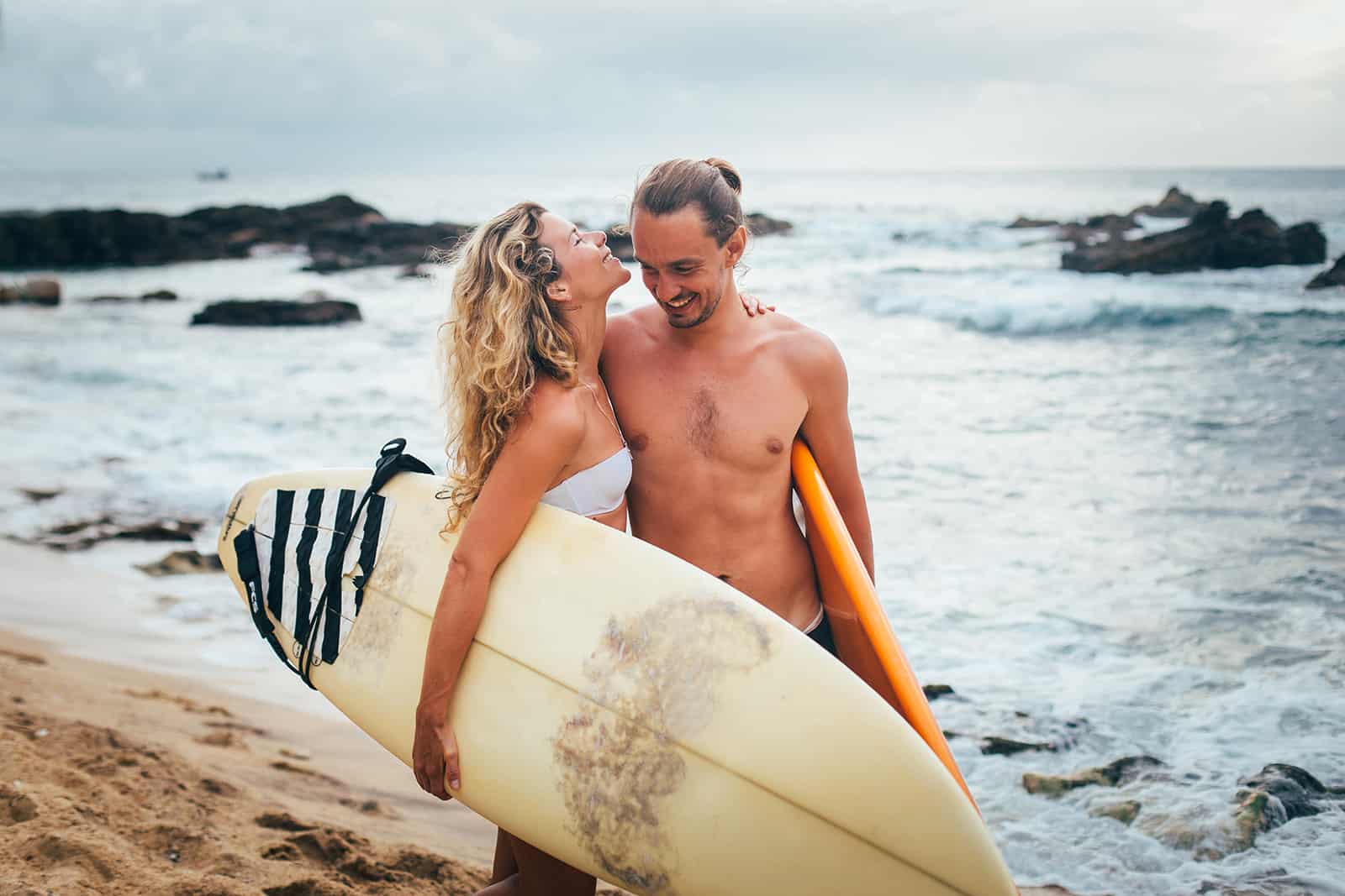 a happy couple carrying surfing boards and walking on the beach