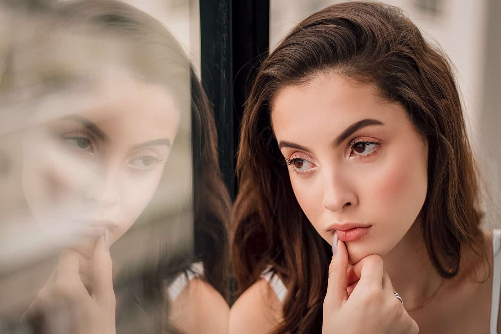 a pensive woman looking through the window and touching lips with thumb