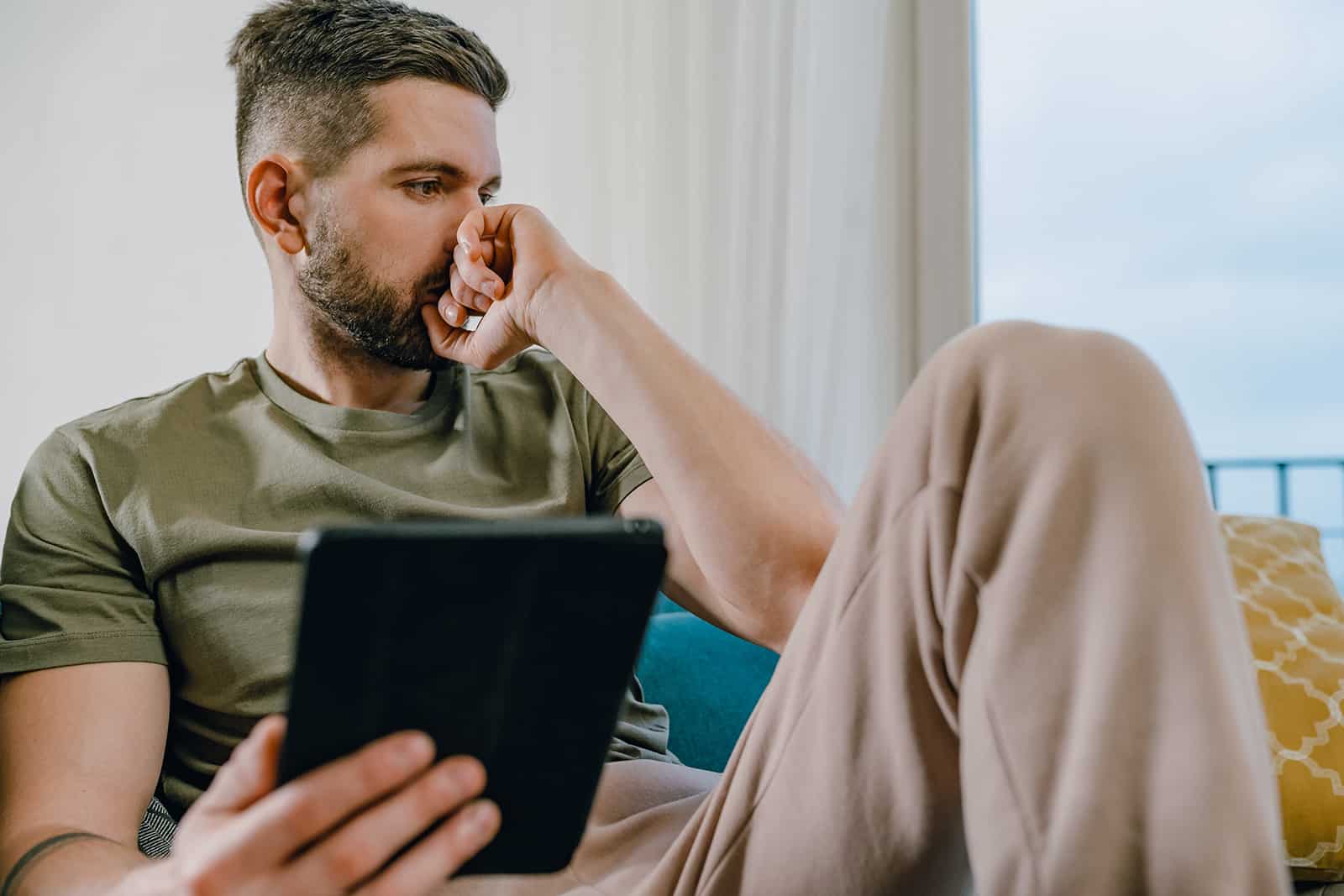 a sad pensive man holding a tablet sitting on the couch at home