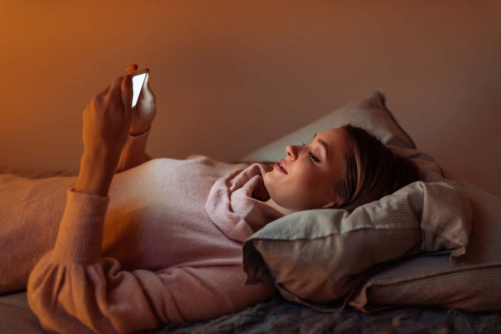 una bionda sorridente si sdraia a letto e utilizza uno smartphone