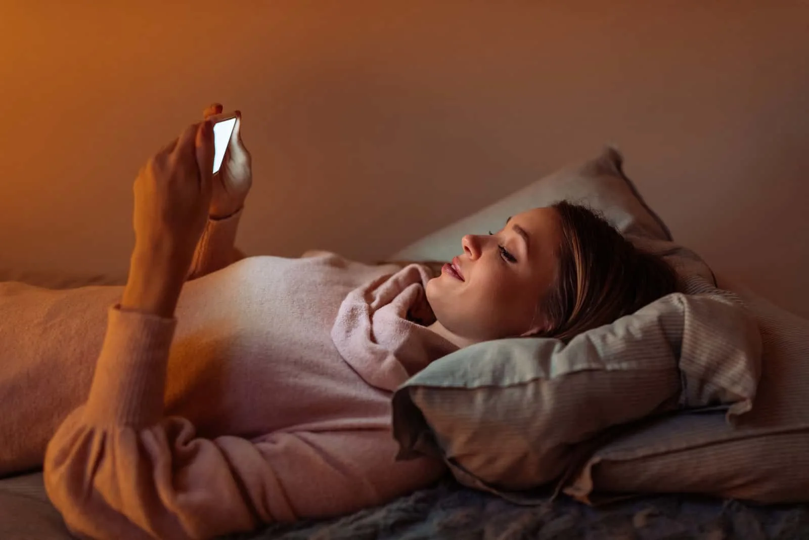 a smiling blonde lies in bed and uses a smartphone