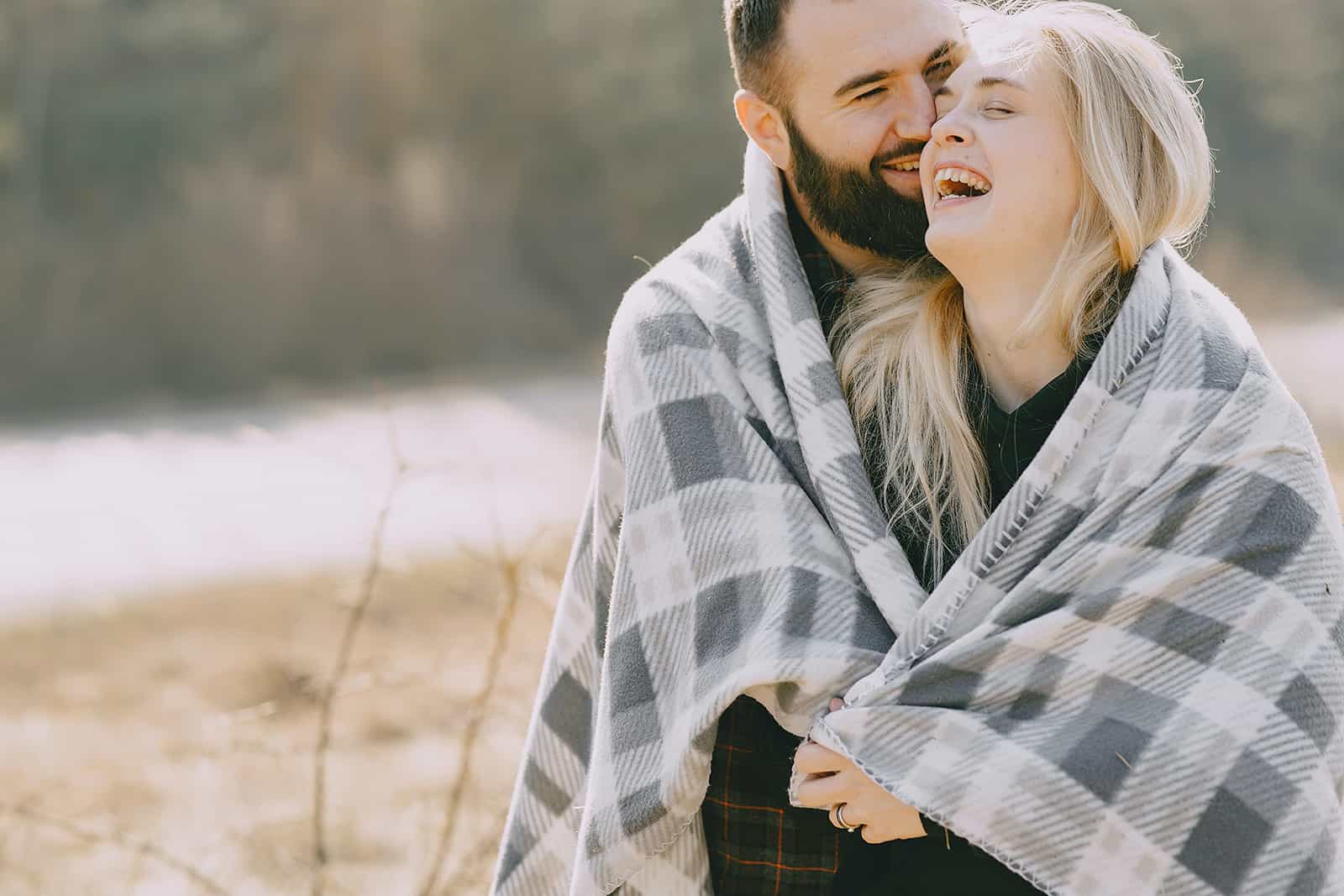 a smiling man embracing his smiling girlfriend while standing outside covered with blanket