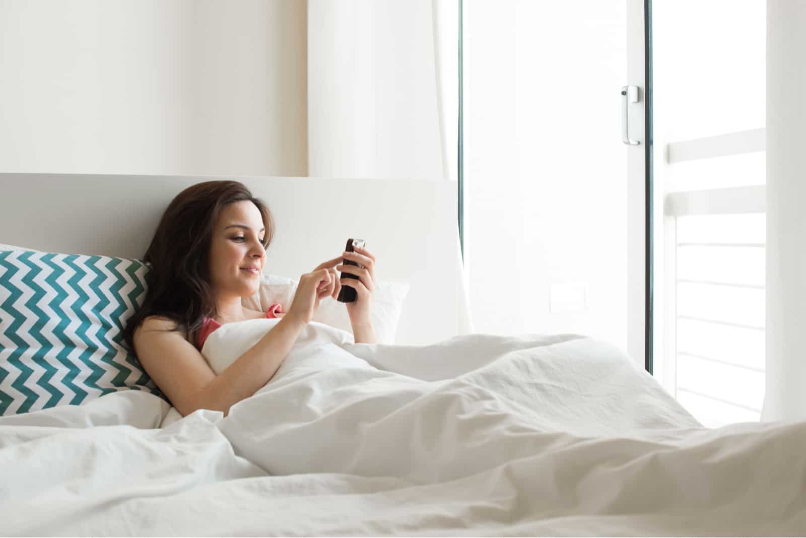 a smiling woman lies in bed and keys on the phone