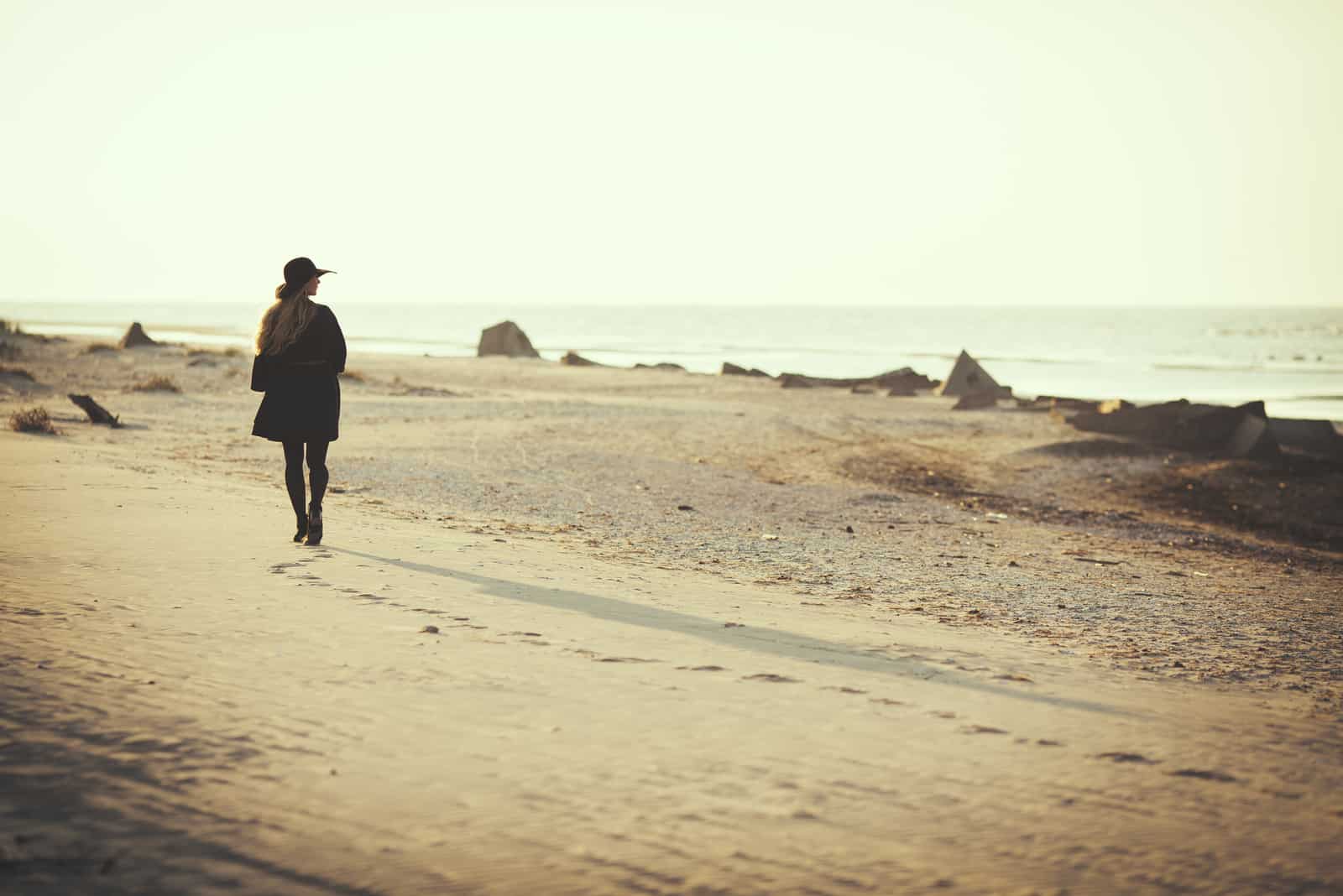 uma mulher com um chapéu preto na cabeça e um casaco preto põe-se na praia