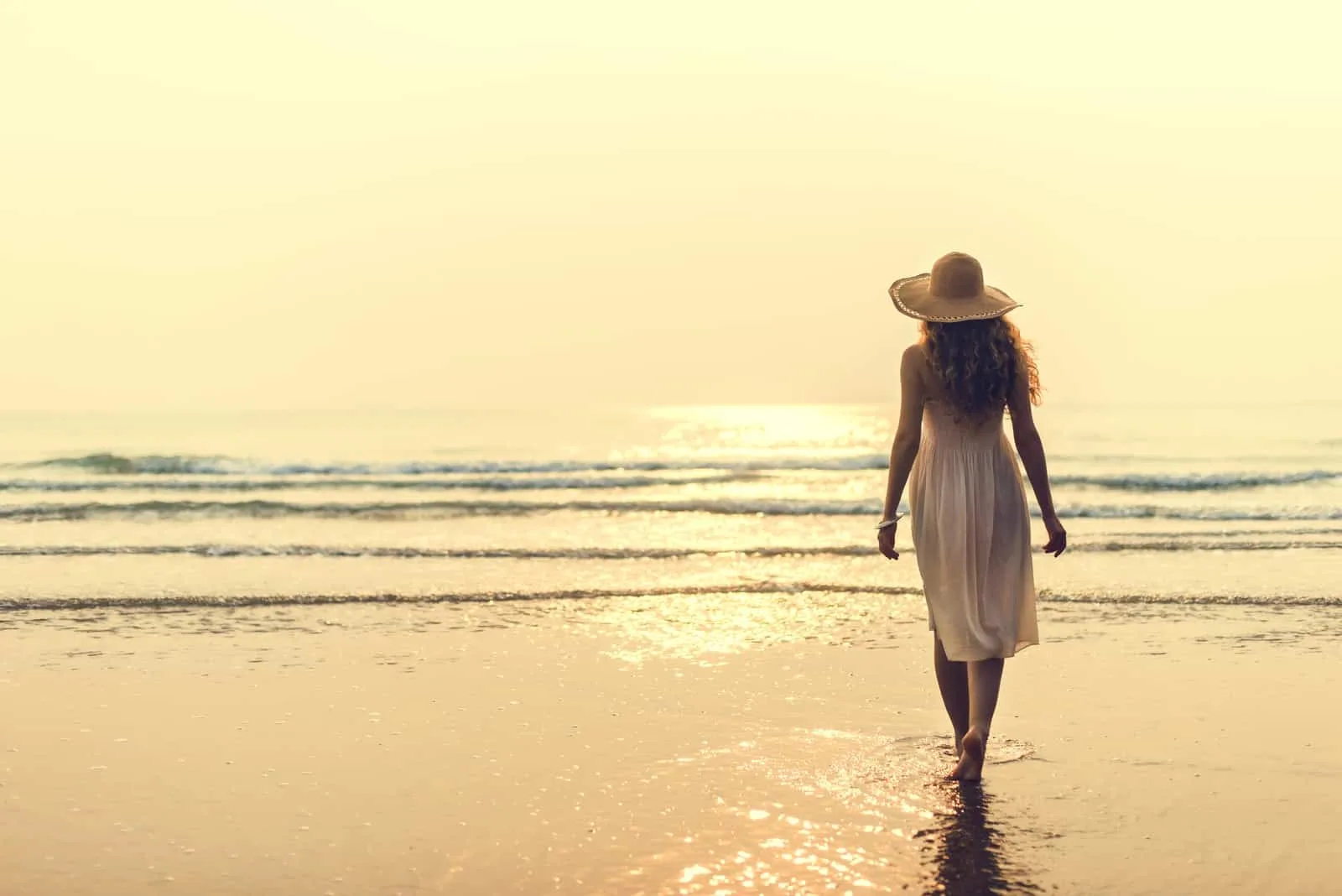 at sunset a barefoot woman in a white dress walks along the beach