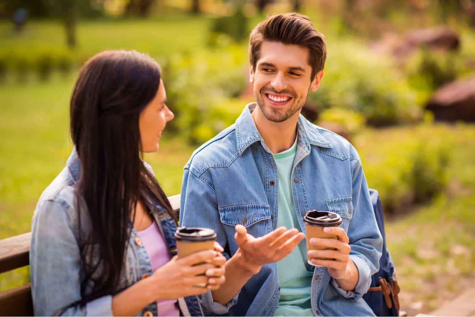 no parque, num banco, um casal sorridente bebe café para levar e conversa