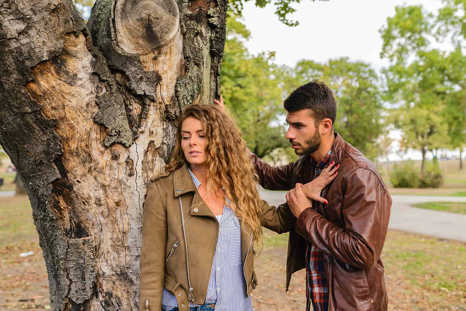 homem a persuadir uma mulher a falar com ele enquanto ela o empurra com a palma da mão para junto da árvore