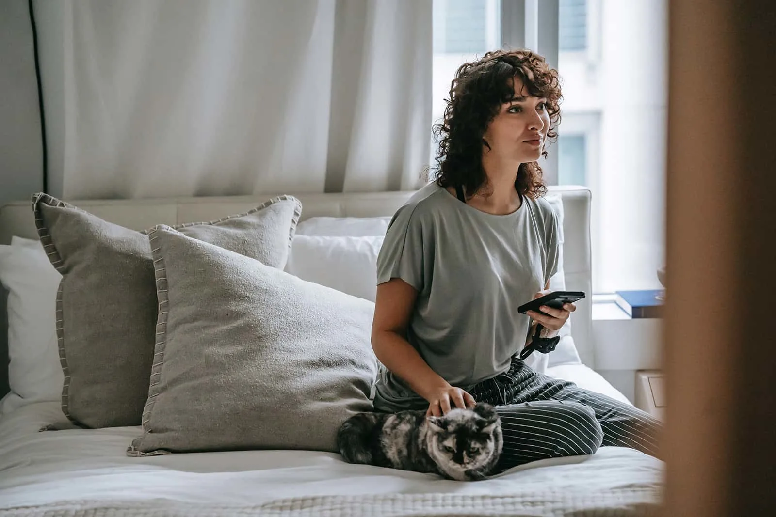 pensive woman holding smartphone while sitting on the bed