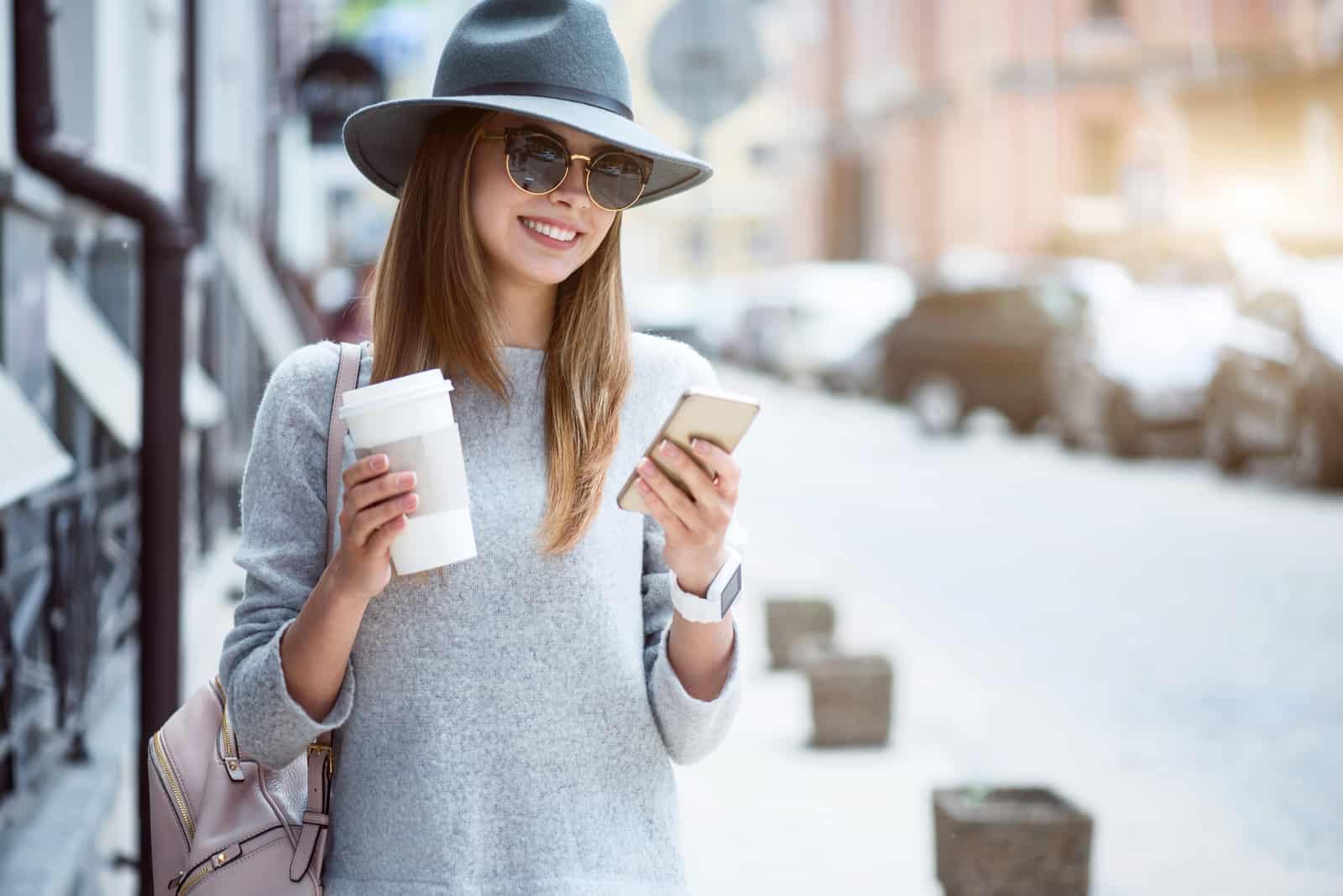 a smiling woman with a hat on her head is standing on the street and a button on the phone