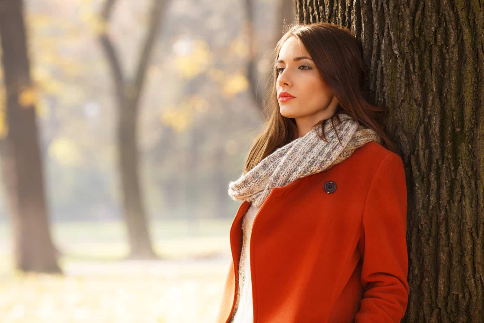 a woman with long brown hair in a red coat stands leaning against a tree
