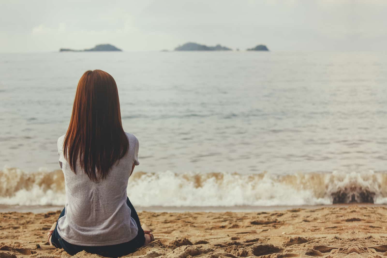 a woman with long brown hair sits on the beach and looks out to sea