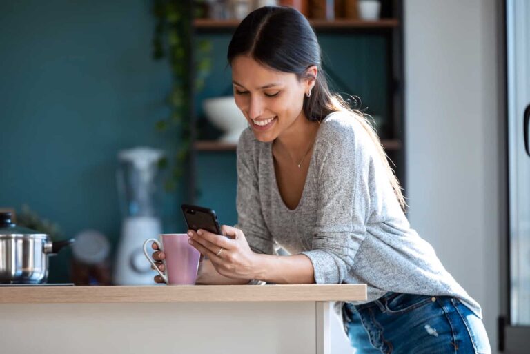a smiling woman standing in the kitchen and pressing a phone