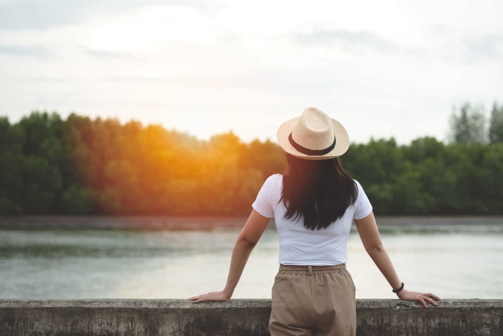 a black-haired woman with a hat on her head stands and looks at the river