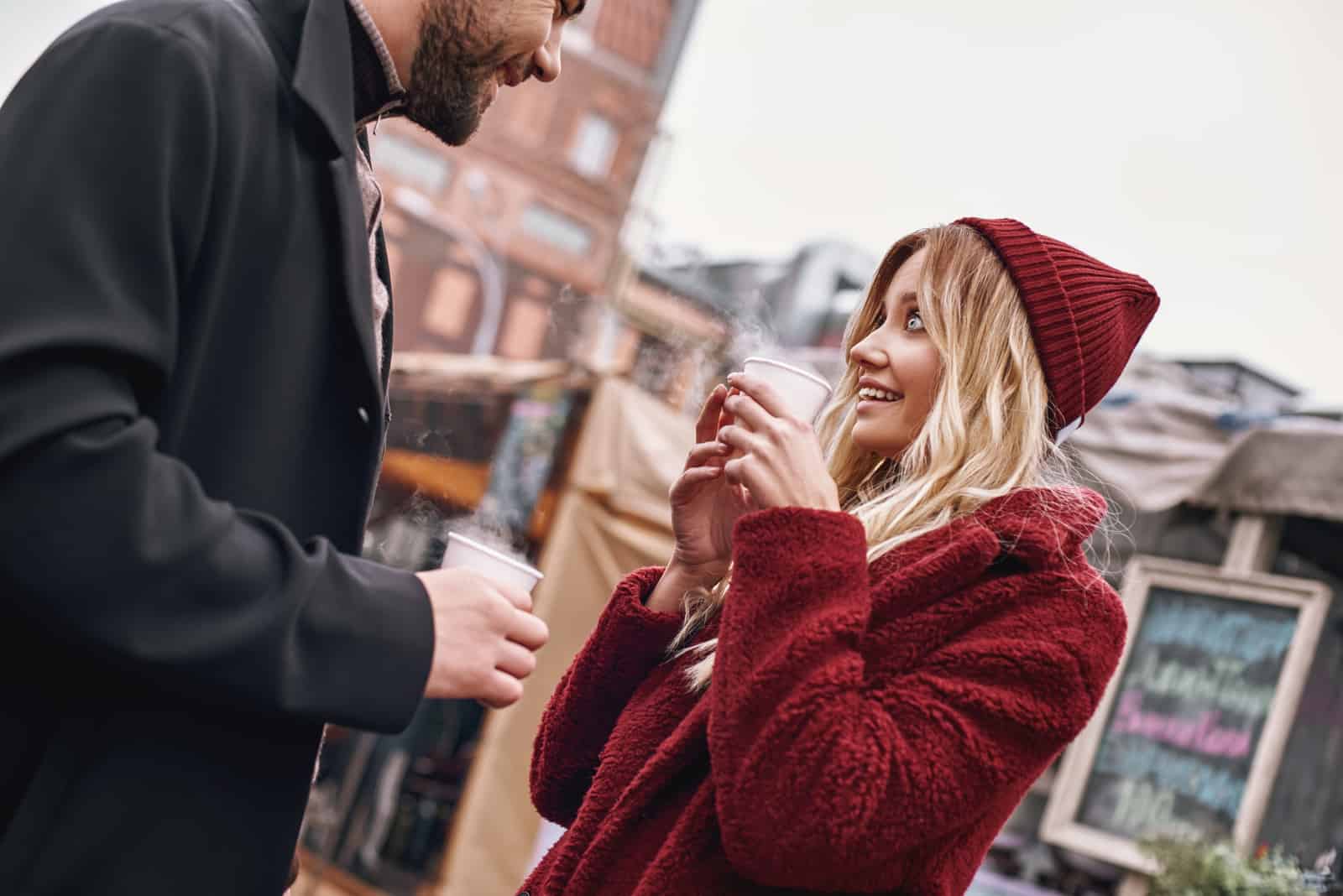 a man and a woman standing outdoors holding coffee in their hands and talking