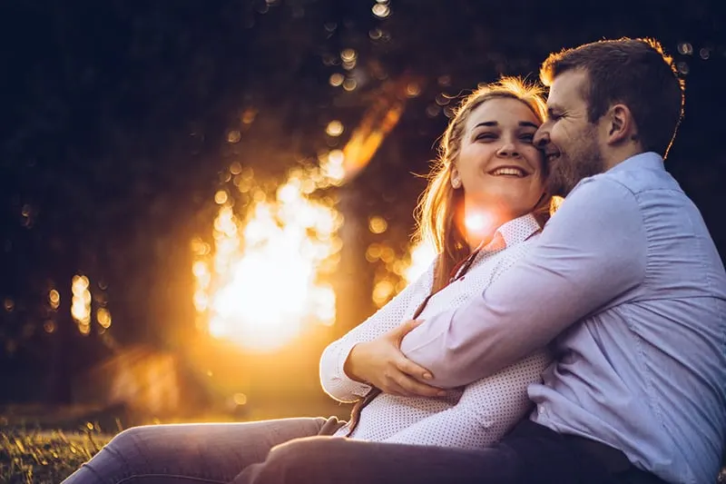 a man hugging smiling woman from the back while sitting on the grass together