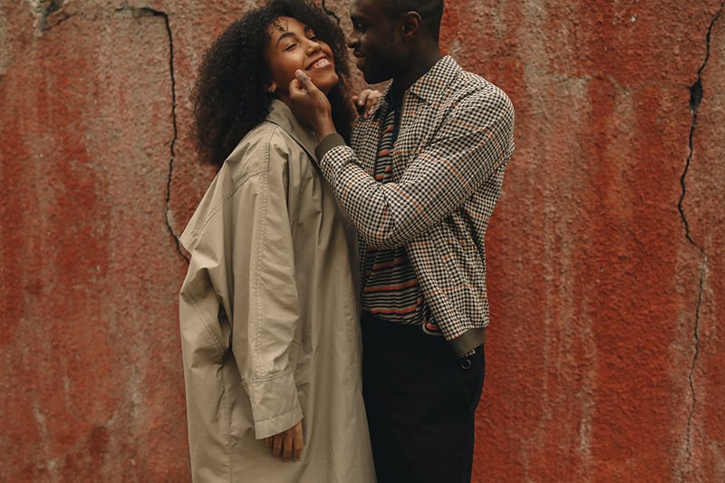 a man touching smiling girlfriend face while standing together near the red wall