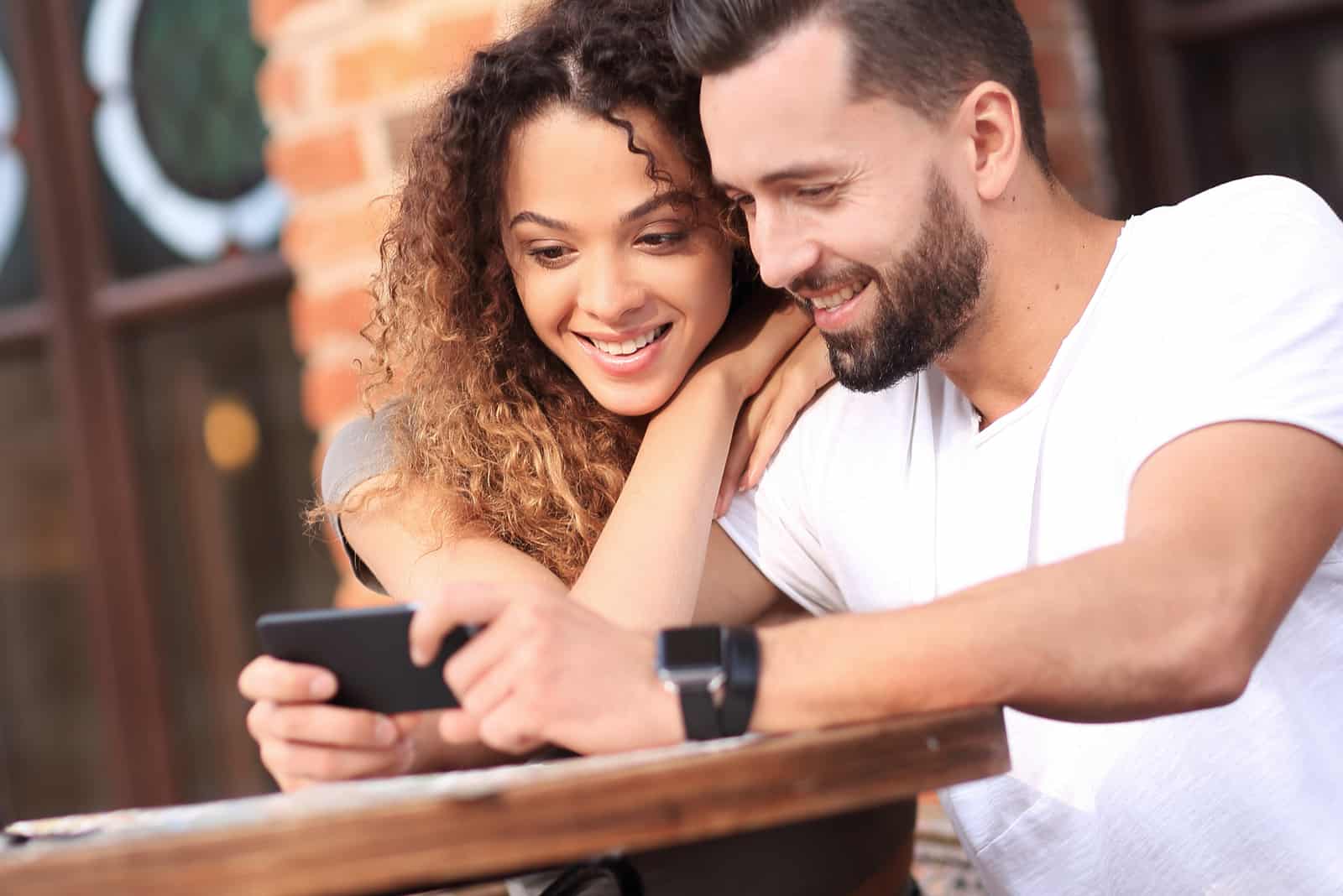 una pareja sonriente sentada a una mesa mirando el teléfono