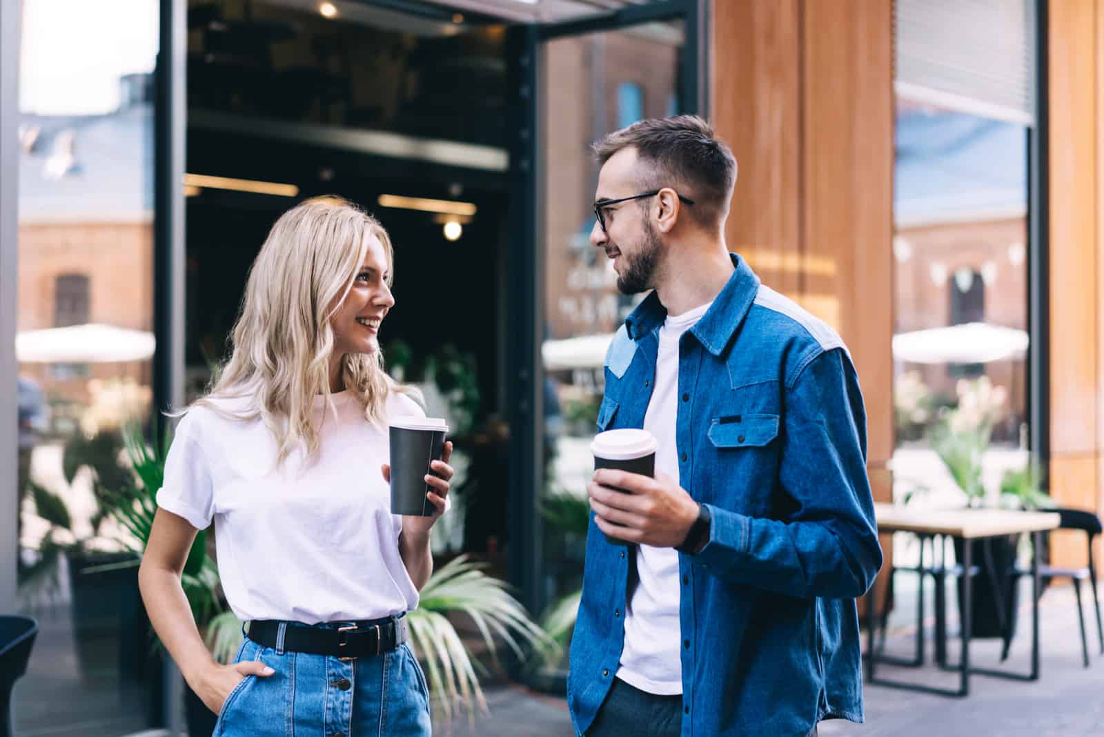 a smiling couple standing in the street holding coffee in their hand and talking
