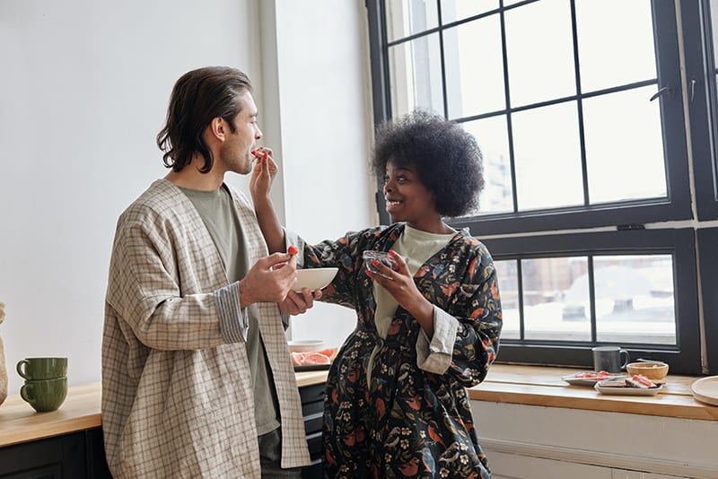 happy woman feeding her boyfriend with strawberries while standing in the kitchen