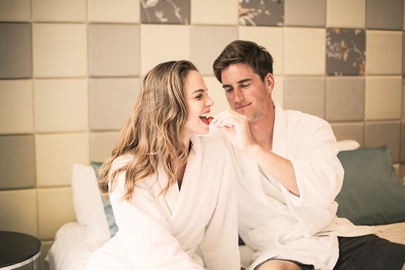 man feeding his girlfriend with strawberry sitting on the bed in bathrobe