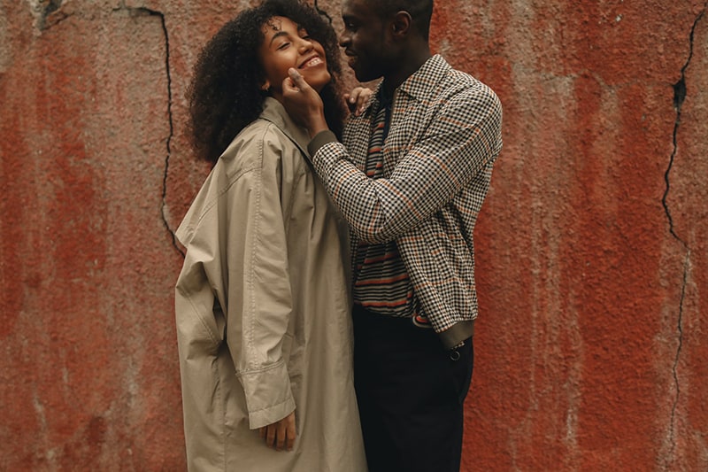 man touching cheek of his smiling girlfriend while standing near the wall