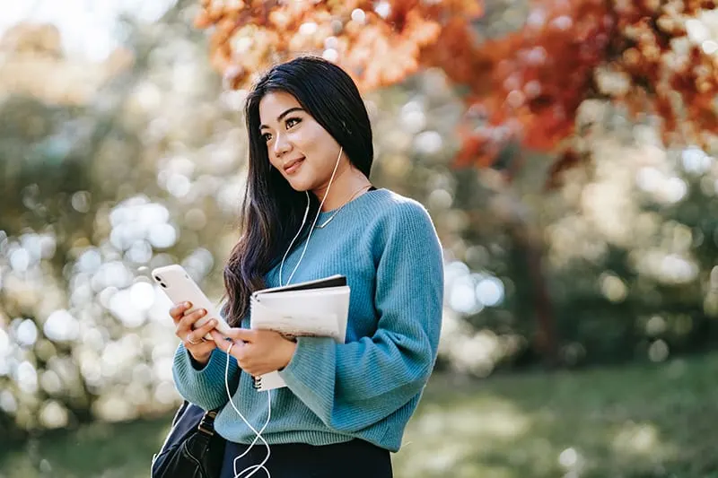 positive young woman listening to music using smartphone and standing in the park