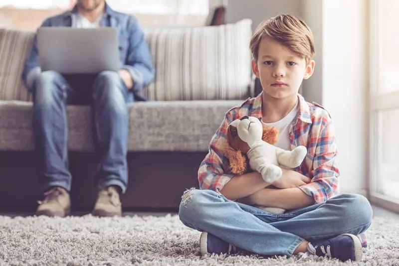 sad little boy sitting on the carpet while his father using laptop