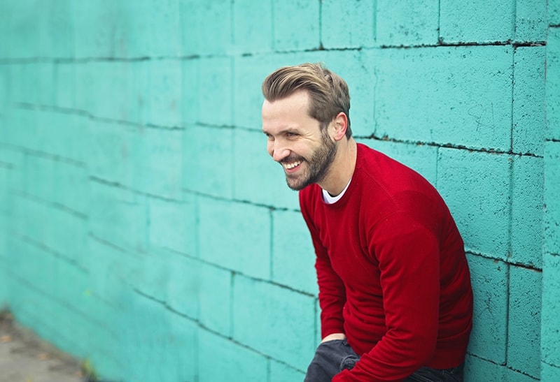 uomo sorridente con maglione rosso appoggiato a un muro blu