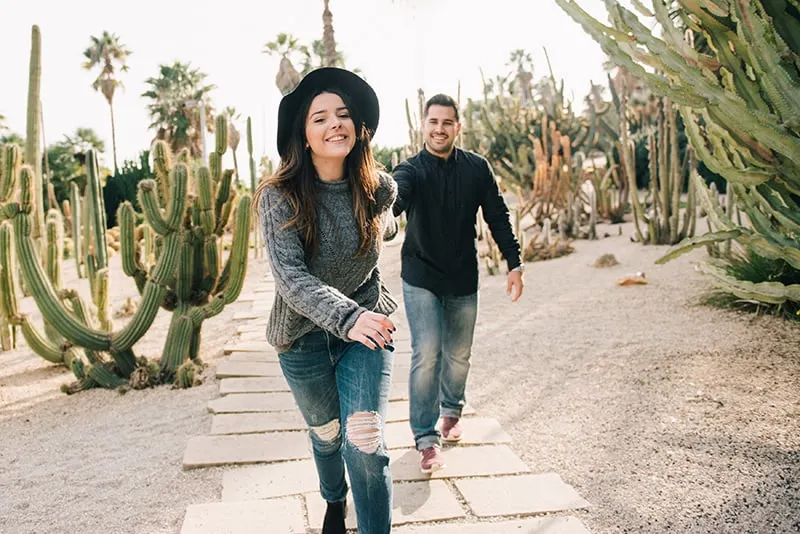 smiling woman pulling her boyfriend for a hand while walking beside cactus field