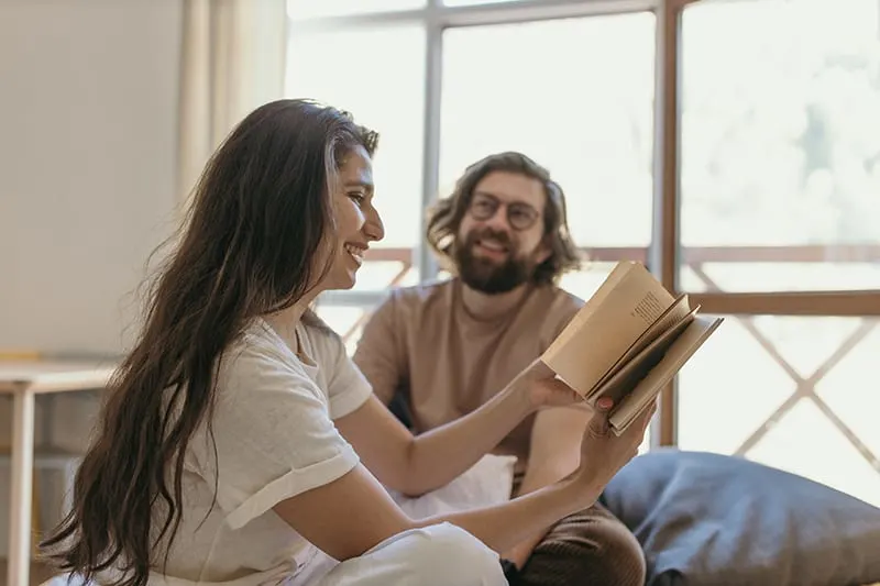 smiling woman reading a book to her boyfriend while sitting on the bed