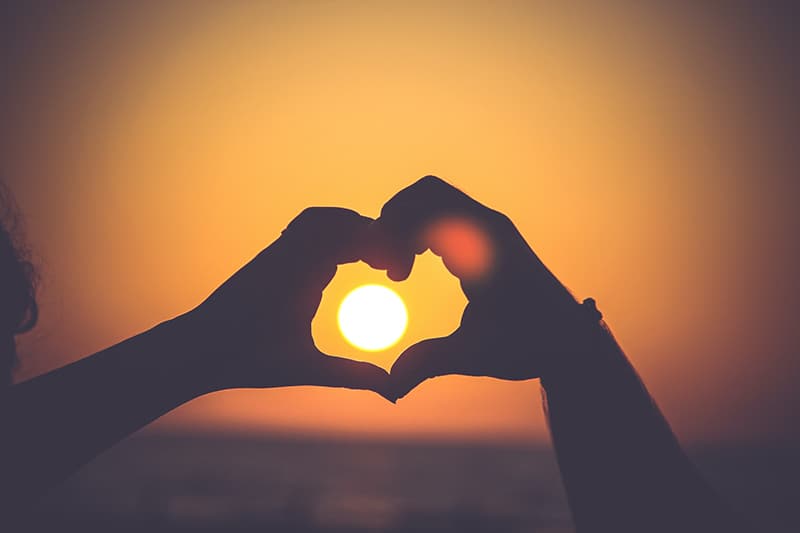 two person forming a heart shape with hands at sunset