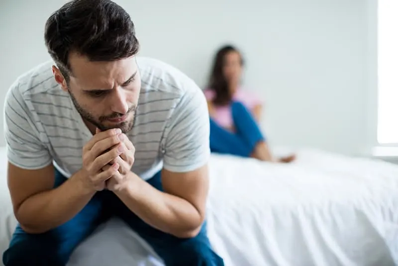 upset man sitting on the edge of the bed ignoring his wife lying on the bed